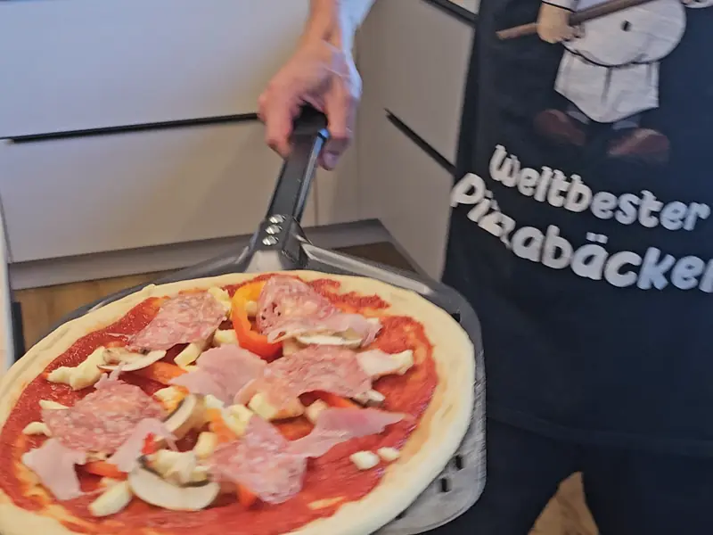 Daniel mit der Schürze „Weltbester Pizzabäcker“ trägt eine belegte Pizza zum Ofen
